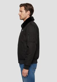 Bomber nero con colletto in pelliccia, caratterizzato da una chiusura frontale con zip, tasche laterali e polsini elasticizzati. Indossato con jeans blu.