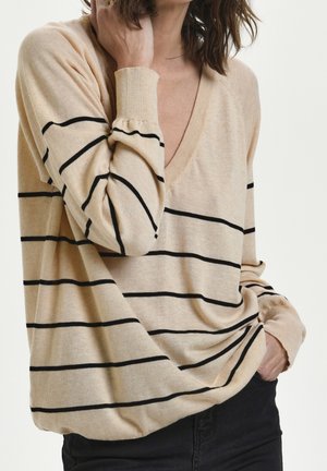 Femme portant un pull beige à col en V avec des rayures horizontales noires, une main touchant le cou, l'autre main dans la poche, assorti à un pantalon noir.