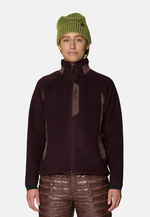 Personne portant un bonnet en maille vert, une veste polaire marron foncé zippée et un pantalon matelassé marron brillant, debout devant un fond uni.