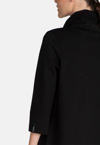 Musta takki, jossa on korkea kaulus ja kolmanneksen pituiset hihat. Sileä pinta, jossa on huomaamaton etiketti hihan lähellä. Yksinkertainen, minimalistinen muotoilu.