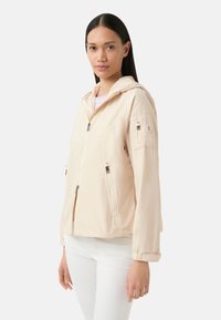 Veste à capuche beige clair avec fermeture éclair sur le devant, poches latérales et un patch sur la manche. Fabriquée en tissu lisse et léger.