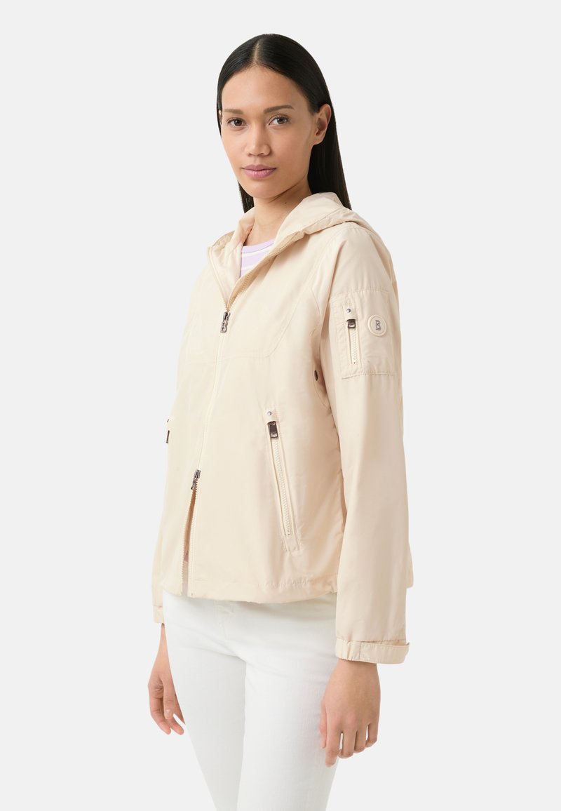 Veste à capuche beige clair avec fermeture éclair sur le devant, poches latérales et un patch sur la manche. Fabriquée en tissu lisse et léger.