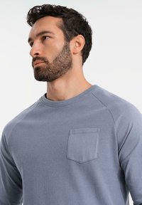 Grijze lange mouwen shirt met een textuurstof, ribbelpatroon en een klein zakje op de linkerborst. Ronde halslijn en raglanmouwen.