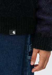 Ranka ilsėdama šalia mėlyno džinsinio kišenės, dėvi juodai-violetinį struktūrizuotą megztinį su matomu Carhartt prekės ženklo etikete.