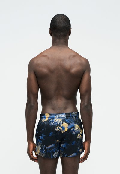 Άνδρας με σκούρο δέρμα που στέκεται με την πλάτη στραμμένη, φοράει σκούρο μαγιό με σχέδια από μπλε και κίτρινους κοραλλιογενείς υφάλους και ψάρια.