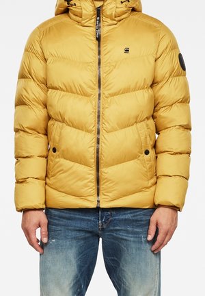 Uomo che indossa una giacca imbottita trapuntata color senape con cerniera frontale e tasche, abbinata a jeans blu slavati.