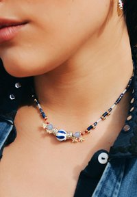 Collier en perles avec une perle centrale blanche rayée de bleu, entourée de perles dorées et colorées, ornée d'accents floraux en or.