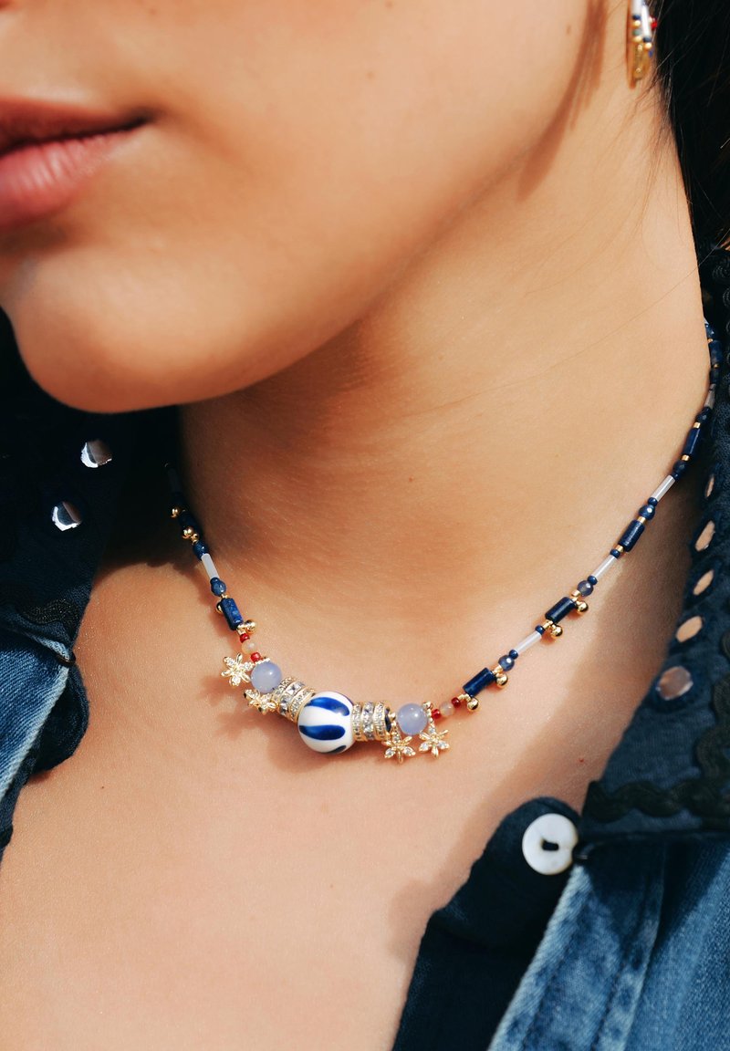 Collier en perles avec une perle centrale blanche rayée de bleu, entourée de perles dorées et colorées, ornée d'accents floraux en or.