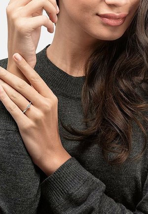 Silberner Ring mit einem zentralen klaren Stein, getragen an einer Hand mit hellbrauner Haut. Sanfte graue Strickpullover im Hintergrund.