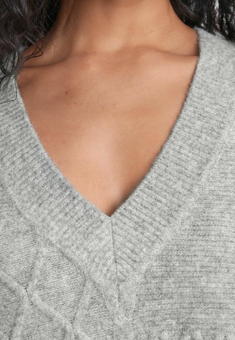 Femme portant un pull léger gris clair à col en V en maille torsadée, montrant le décolleté et la zone de la clavicule, avec des cheveux bouclés foncés partiellement visibles.