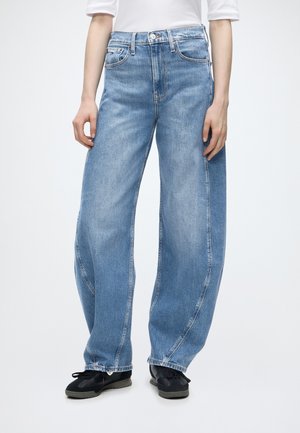 Persoon draagt loszittende lichtblauwe jeans met hoge taille, gecombineerd met zwarte schoenen en een wit shirt met lange mouwen, staand tegen een witte achtergrond.
