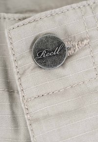Knopf aus beigem Stoff, mit einer metallic grauen Oberfläche und eingraviertem Text "Reell." Strukturiert mit Stichdetails in der Nähe.