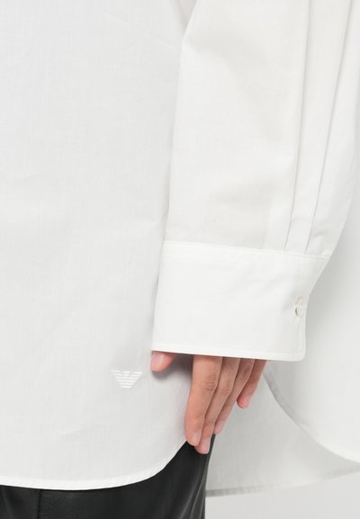 Biela košeľa s dlhým rukávom a hladkou textúrou, s gombíkovým manžetou a malým výšivkovým logom blízko spodného lemu. Detailný záber ruky položené na látke.