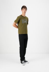 Olivgrünes Baumwoll-T-Shirt mit dem Schriftzug "GAP" in Schwarz und Weiß. Kombiniert mit schwarzen Jeans und schwarz-weißen Sneakern, stehend vor einem schlichten Hintergrund.