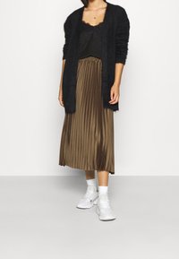 Schwarze, flauschige Strickjacke über einem schwarzen Spitzenoberteil, kombiniert mit einem plissierten, khakifarbenen Midirock und weißen Turnschuhen mit Knöchelsocken. Einfaches, lässiges Outfit.