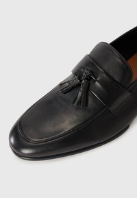 Mocassin en cuir noir avec un bout arrondi et des franges décoratives cousues sur le dessus, sur un fond clair et uni.