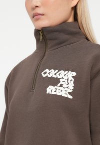 Detailný záber osoby v hnedej mikine na zips s bielym nápisom "COLOURFUL REBEL" na hrudi a vysokým golierom.