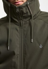 Olivengrøn zip-jakke med høj krave, snoretræk i hætten, sølv lynlås og subtilt logo detalje på brystet.