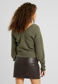 Kobieta z kręconymi włosami, ubrana w zielony sweter z prążkowanej tkaniny oraz mini spódnicę z ciemno-brązowej skóry, stoi bokiem, z widoczną plecami.