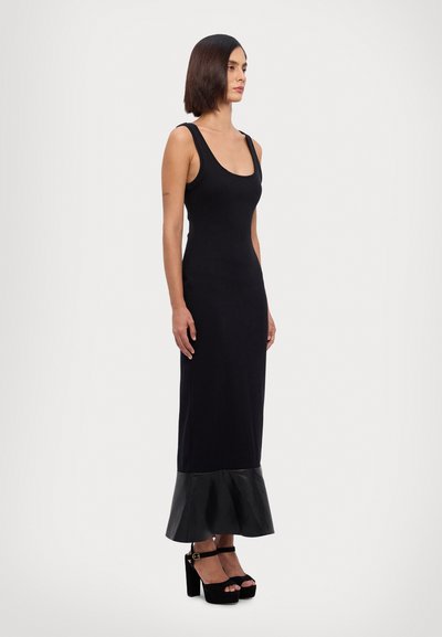 Robe maxi sans manches noire avec une texture côtelée, présentant un ourlet en cuir évasé. Le modèle porte des sandales à plateforme noires et chunky.