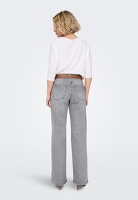 Graue Jeans mit weitem Bein, hoher Taille, detaillierter Naht und zwei Gesäßtaschen; kombiniert mit einem weißen Langarmshirt und braunem Gürtel.