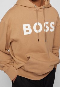 Persona con una sudadera color beige con el grande texto blanco "BOSS" en el pecho, manos en el bolsillo delantero, combinada con pantalones negros.