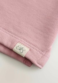 Lyserød bomuldsstof foldet med en syet kant, prydet med et lille stofmærke med blomstermotiv. Teksturen fremstår blød og glat.