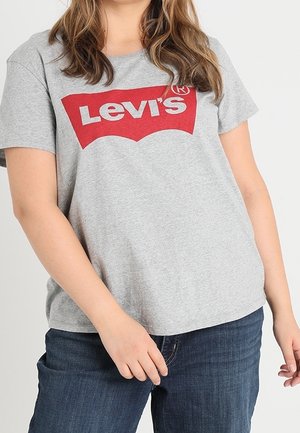 Person iført en grå Levi’s T-shirt med rød logo og blå jeans, stående mod en ensfarvet lys baggrund.