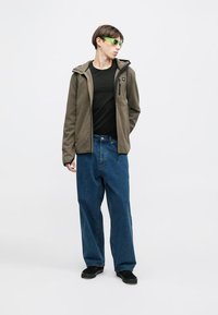 Ελαιόχρωμο φερμουάρ μπουφάν, μαύρο t-shirt, χαλαρά μπλε τζιν παντελόνια και μαύρα παπούτσια. Αξεσουάρ περιλαμβάνουν γυαλιά ηλίου με πράσινο φίλτρο.