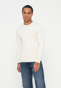 Joven vestido con un suéter blanco roto con el logo "GUESS" en relieve y jeans azules, de pie frente a un fondo blanco liso.