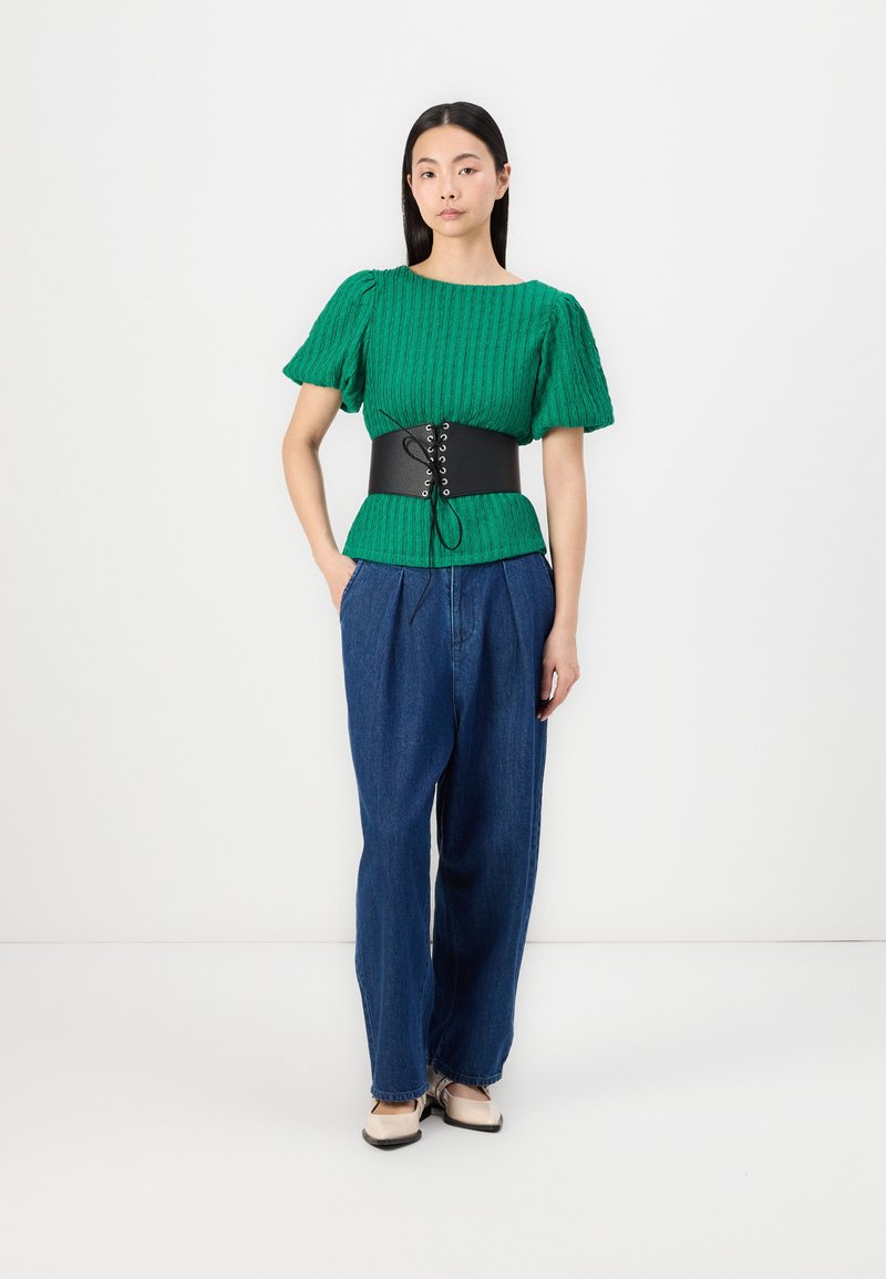 Zielony prążkowany sweter z bufkami, czarny pas koronkowy, niebieskie dżinsy z wysokim stanem i szerokimi nogawkami oraz beżowe buty z czarnymi akcentami.