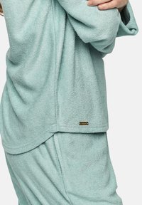 Ensemble de loungewear en tissu éponge bleu clair, présentant une texture douce, une coupe décontractée et une petite étiquette logo en bas à gauche du haut.