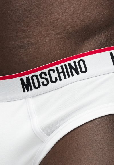 Λευκά εσώρουχα με κόκκινη και λευκή μέση που φέρει την επωνυμία "MOSCHINO", φορεμένα πάνω σε σκούρο δέρμα, κοντινή λήψη στη περιοχή του ισχίου και της μέσης.