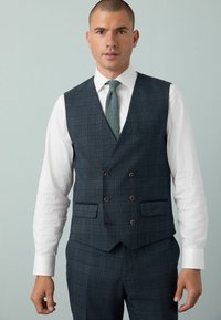 Homme portant un gilet à carreaux gris foncé croisé et un pantalon assorti, avec une chemise blanche et une cravate en texture verte, contre un fond gris clair.