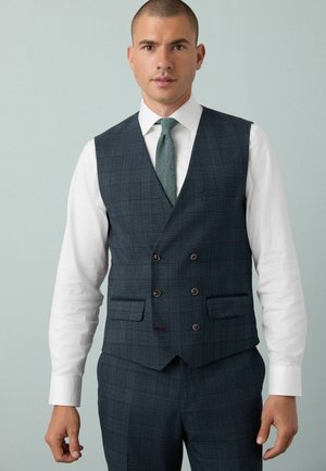 Man wearing dark gray checked double-breasted vest and matching trousers with white shirt and green textured tie against light gray background.