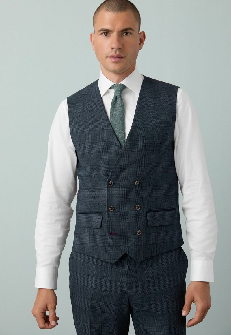 Homme portant un gilet à carreaux gris foncé croisé et un pantalon assorti, avec une chemise blanche et une cravate en texture verte, contre un fond gris clair.