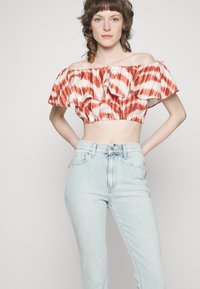 Top cropped com ombros à mostra e decote cheio de volantes, em padrão tie-dye vermelho e branco; combinado com jeans de cintura alta em azul claro.