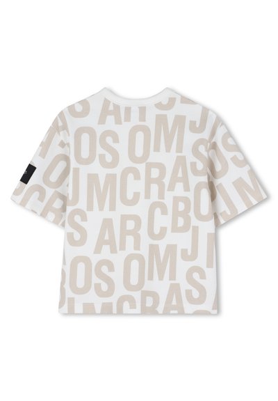 T-shirt blanc à manches courtes avec de grandes lettres beiges imprimées de manière aléatoire sur toute la surface et une petite étiquette noire sur la manche gauche.