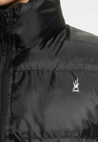 Black puffer jacket with a high collar, featuring a front zipper and a small white logo on the chest. Textured nylon fabric.