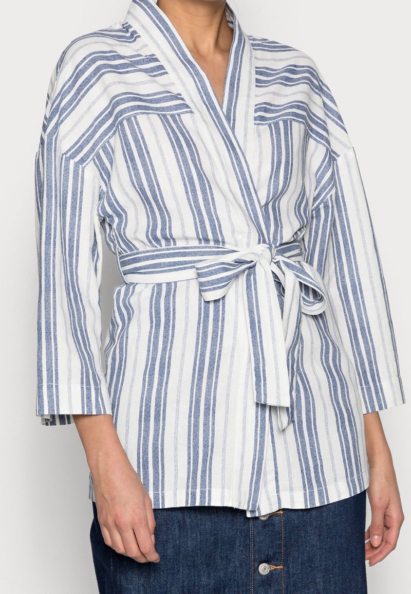 Buste portant une veste portefeuille à rayures verticales blanches et bleues avec des manches trois-quarts nouée à la taille sur une jupe bleue foncée boutonnée.
