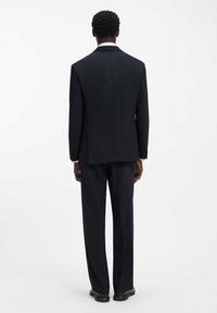 Czarny garnitur na miarę o wyrafinowanym fasonie, z marynarką na jedno zapięcie i spodniami o prostych nogawkach. Gładka tkanina, minimalistyczne zdobienia.