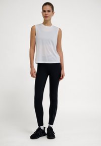 Vit ärmlös topp med rund halsringning, kombinerad med svarta leggings med ett subtilt diagonalt mönster och svarta träningsskor.