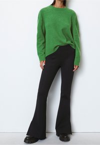 Vihreä neulepaita pitkillä hihoilla ja pyöreällä kauluksella, yhdistetty mustiin leveälahkeisiin housuihin. Mustat paksupohja sneakersit näkyvät alhaalla.