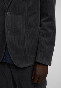 Blazer in velluto a coste di colore grigio scuro con texture a coste verticali, chiusura a un bottone e tasche laterali. I polsini presentano un dettaglio di cucitura sottile.