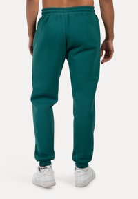 Pantalones de chándal en color verde azulado con cintura elástica, perneras ajustadas y puños acanalados, combinados con zapatillas blancas. Tela suave, con detalles mínimos.