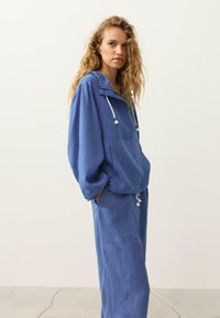 Jeune femme aux cheveux blonds bouclés portant un pull à capuche bleu ample et un pantalon large assorti, debout les mains dans les poches sur un fond uni.