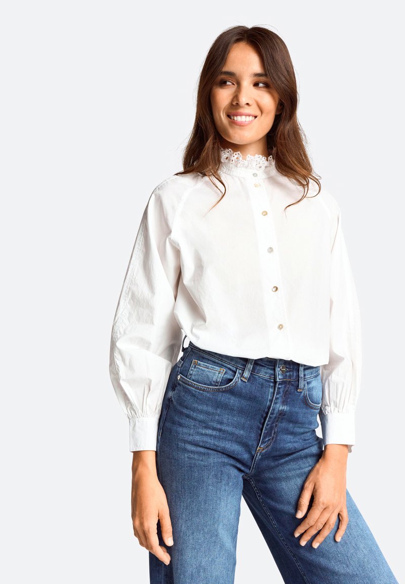 Weißes Hemd mit hohem Spitzenkragen, langen Puffärmeln und subtiler Textur, kombiniert mit hochgeschnittenen blauen Jeans.