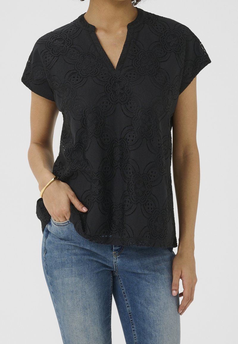 Femme portant un chemisier noir texturé à manches courtes avec des motifs floraux et un jean bleu, main dans la poche, et un bracelet en or au poignet gauche.