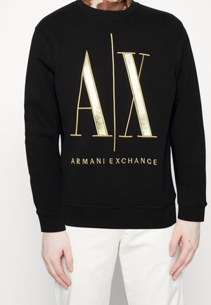 Sweat-shirt noir à manches longues avec grand logo doré "A|X" et texte "Armani Exchange", porté avec un pantalon blanc.