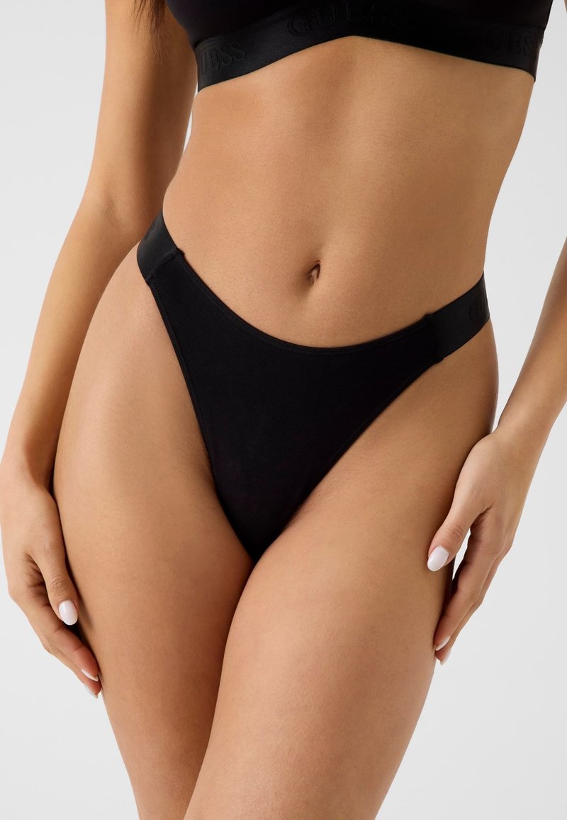 Femme portant des sous-vêtements noirs taille haute et un soutien-gorge de sport assorti, montrant le ventre et les mains avec du vernis à ongles blanc.